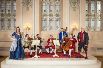 Barockensemble in historischen Kostümen bei einem Konzert in einem festlichen Saal mit Kronleuchter und Streichinstrumenten.