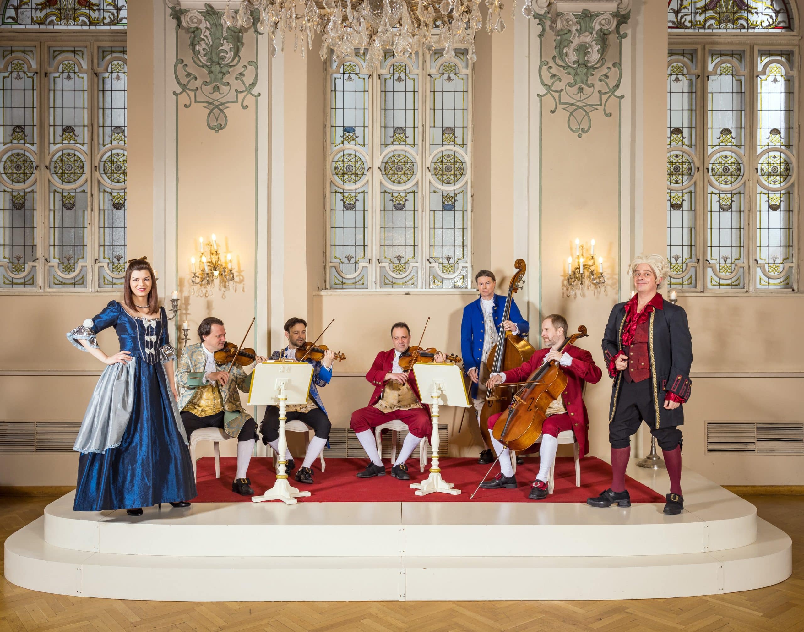 Barockensemble in historischen Kostümen bei einem Konzert in einem festlichen Saal mit Kronleuchter und Streichinstrumenten.